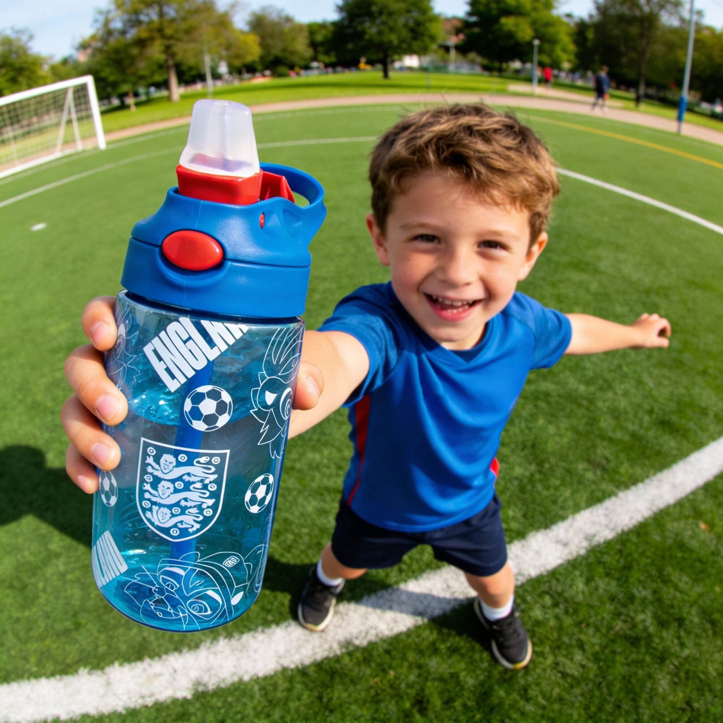 YOUNG BOY HOLDING OFFICIALLY LICENSED ENGLAND WATER BOOTTLE WITH KID FRIENDLY PRINT IN BLUE ON A GRASS FOOTBALL PITCH