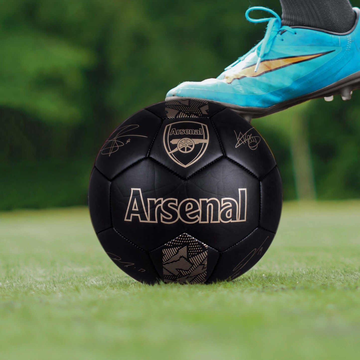 Black Arsenal ball with gold metallic logo and signatures on a grass football pitch with a blue football boot on top of the ball.