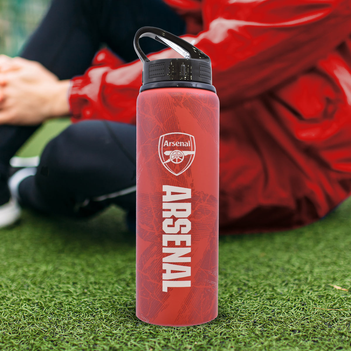 Arsenal F.C. Aluminium Bottle in red and black on a grass football pitch background with a person sat next to it wearing red