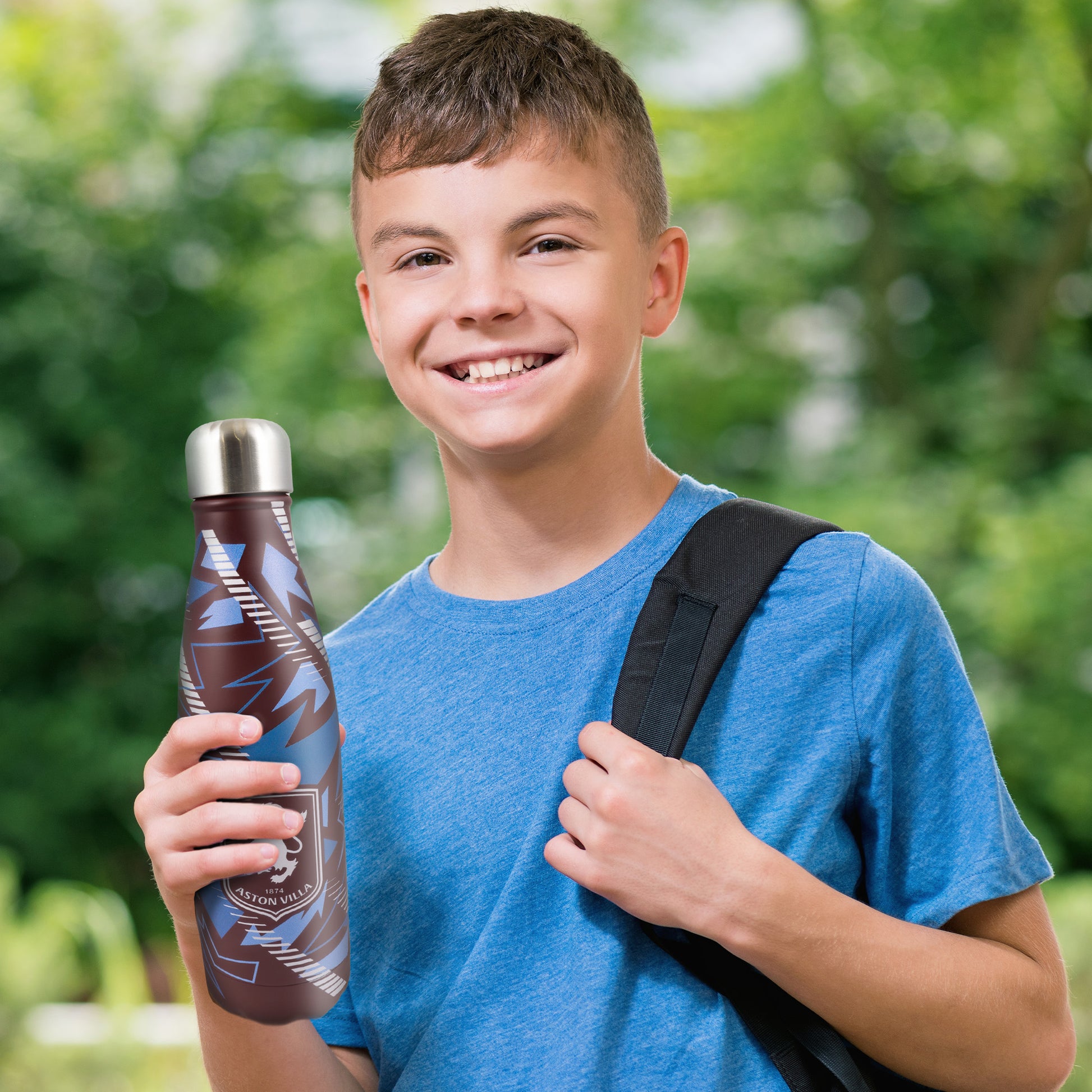 A child smiling while holding the Officially licensed Aston Villa thermal water bottle by Hy-Pro Sports. Background is a green park like setting 