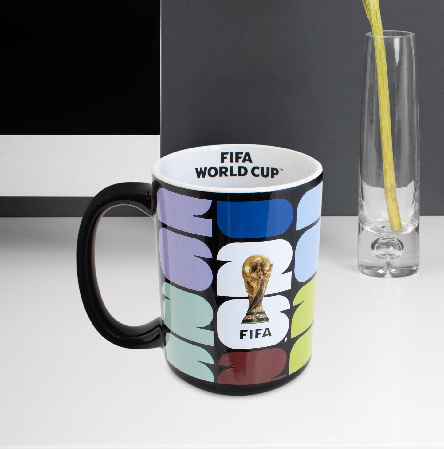 OFFICIAL FIFA WORLD CUP 2026 CERAMIC MUG IN BLACK WITH 26 BRANDING BY HY-PRO ON A DESK WITH A COMPUTER SCREEN IN  THE BACKGROUND