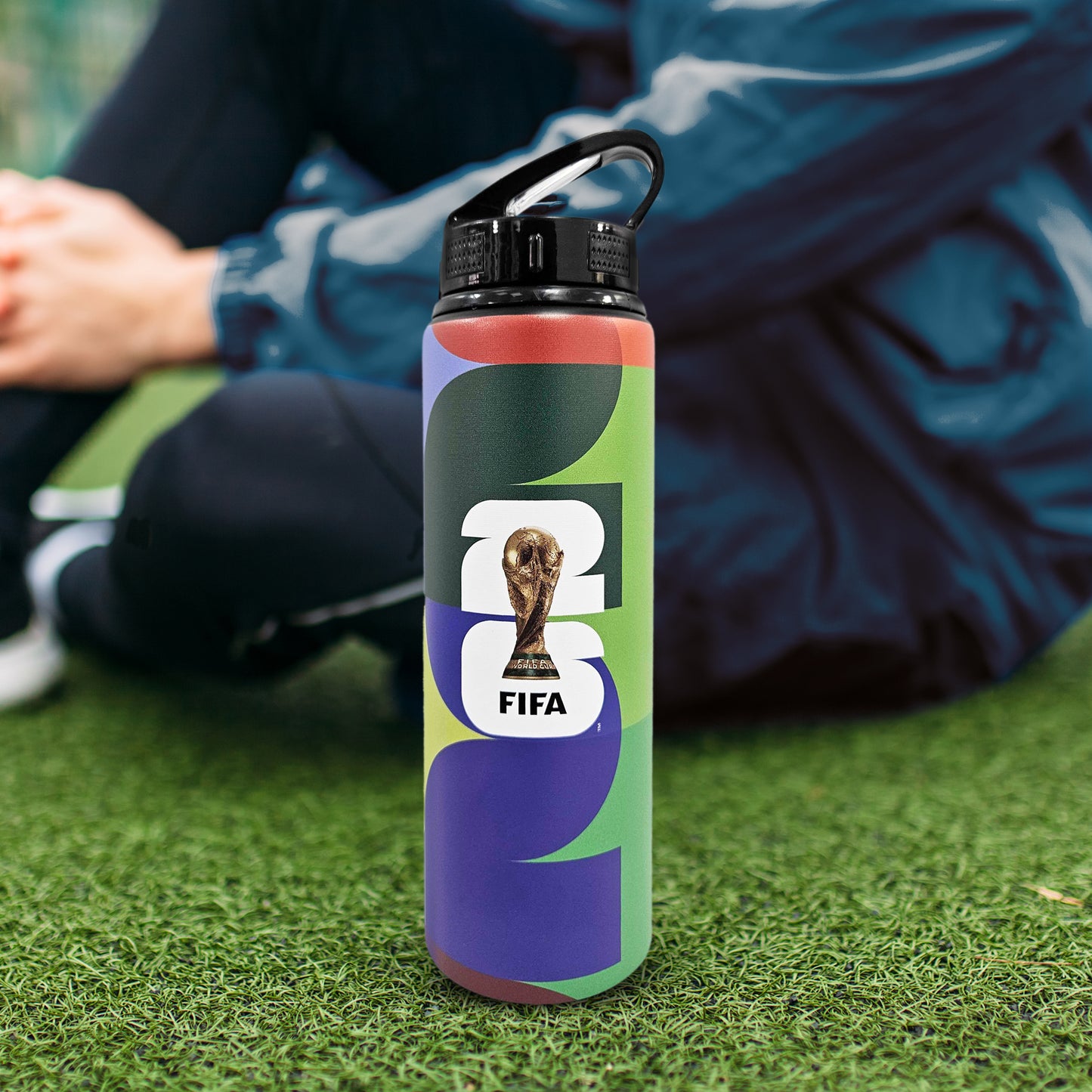 FIFA WORLD CUP 2026 750ML ALUMINIUM WATER BOTTLE BY HY-PRO SPORTS ON A GRASS FIELD WITH A PLAYER SAT NEXT TO IT