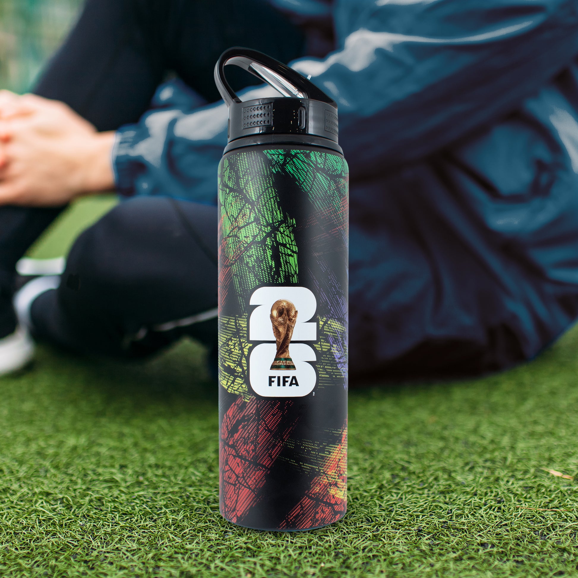 FIFA WORLD CUP 2026 750ML ALUMINIUM WATER BOTTLE BY HY-PRO SPORTS ON A GRASS FIELD WITH A PLAYER SAT ON THE FLOOR BEHIND IT