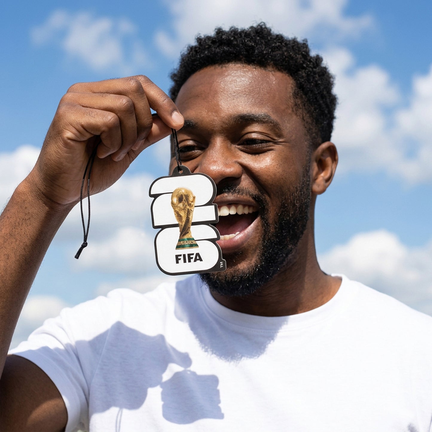 OFFICIALLY LICENSED FIFA WORLD CUP 2026 AIR FRESHNER IN BLACK AND WHITE BEING HELD BY A SMILING FOOTBALL FAN WITH A BLUE SKY IN THE BACKGROUND
