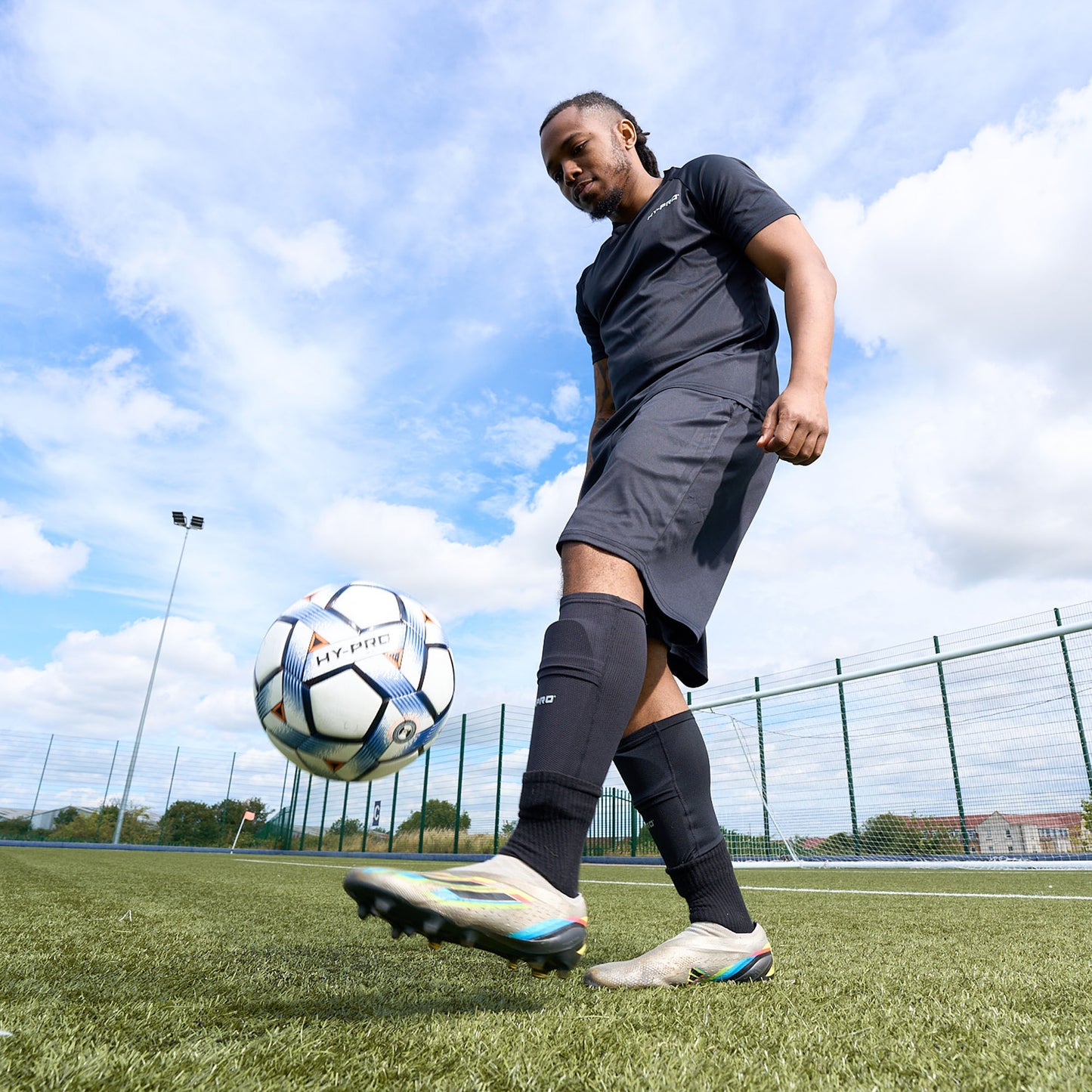 Side view of Hy-Pro Sports Shin Pads being worn by a football player kicking a football into the air on an Astroturf pitch