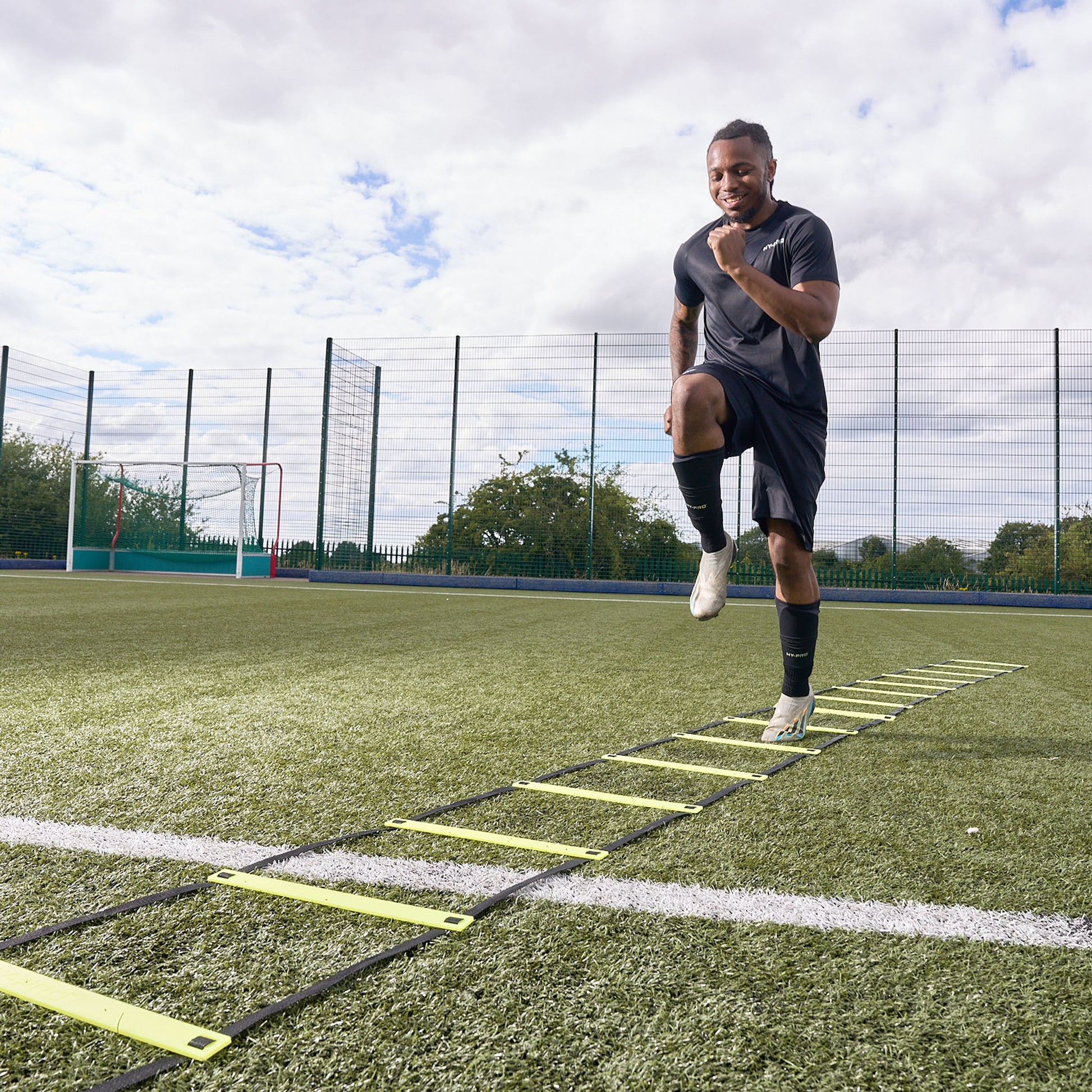 Smiling footballer uses the Hy-Pro Sports yellow agility ladder on an Astroturf football pitch to train agility 